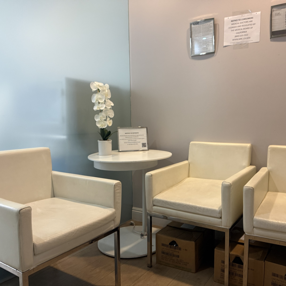 Three white chairs surround a small round table with a white orchid in a pot; cardboard boxes and notices are visible on the wall behind.