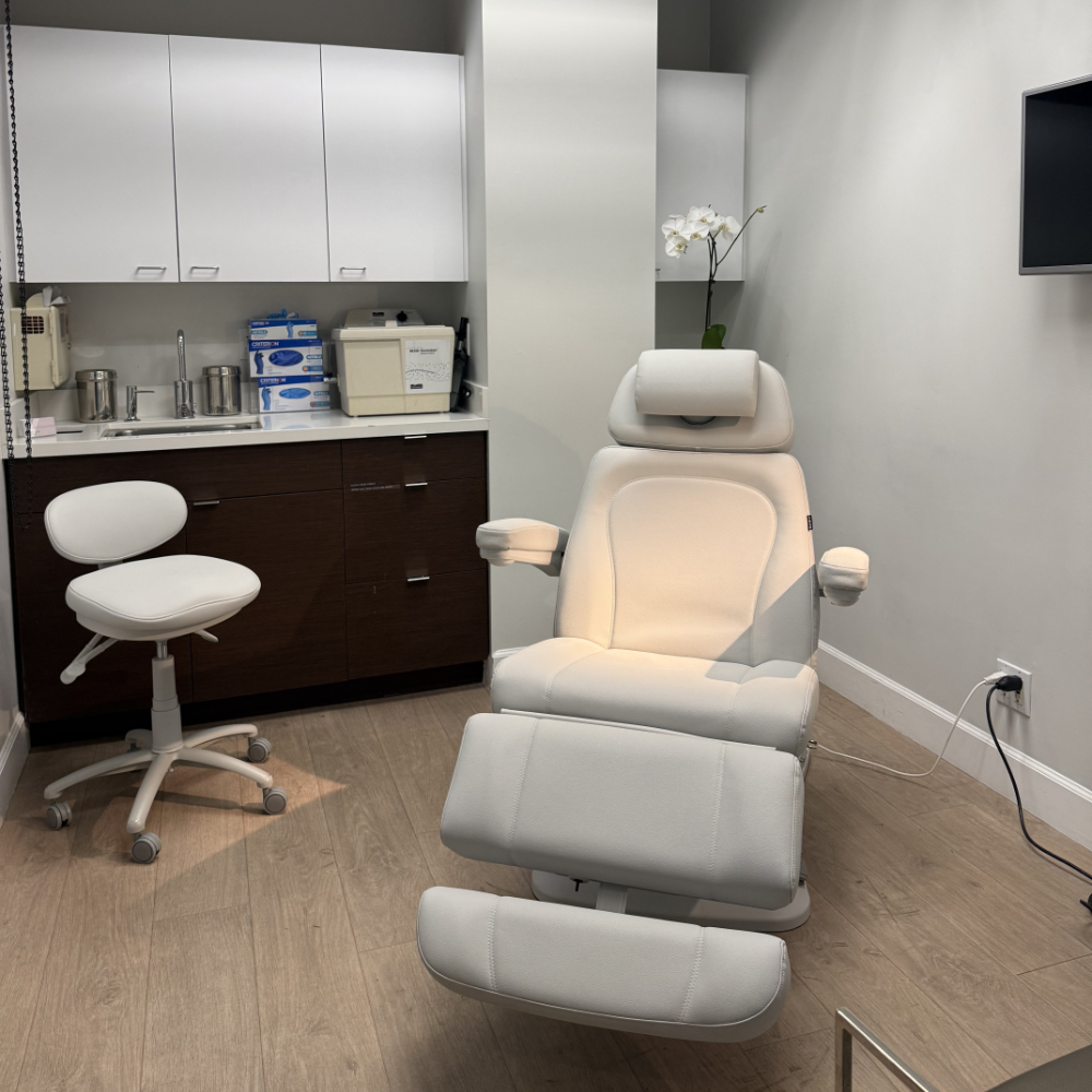A modern medical or spa treatment room with a white reclining chair, stool, cabinets, countertop supplies, and an orchid on the counter.