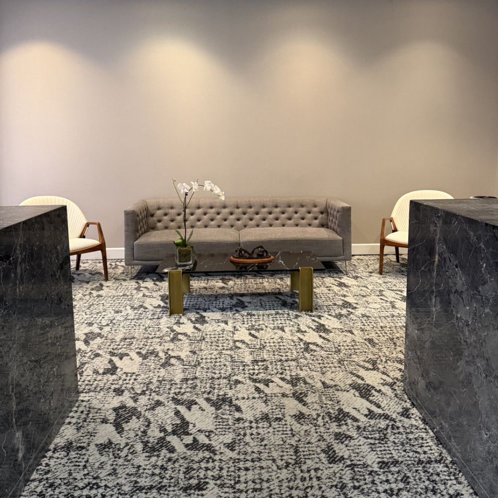 A modern lounge area with a gray sofa, two white armchairs, a marble coffee table with a plant and decorative items, and patterned carpeting.