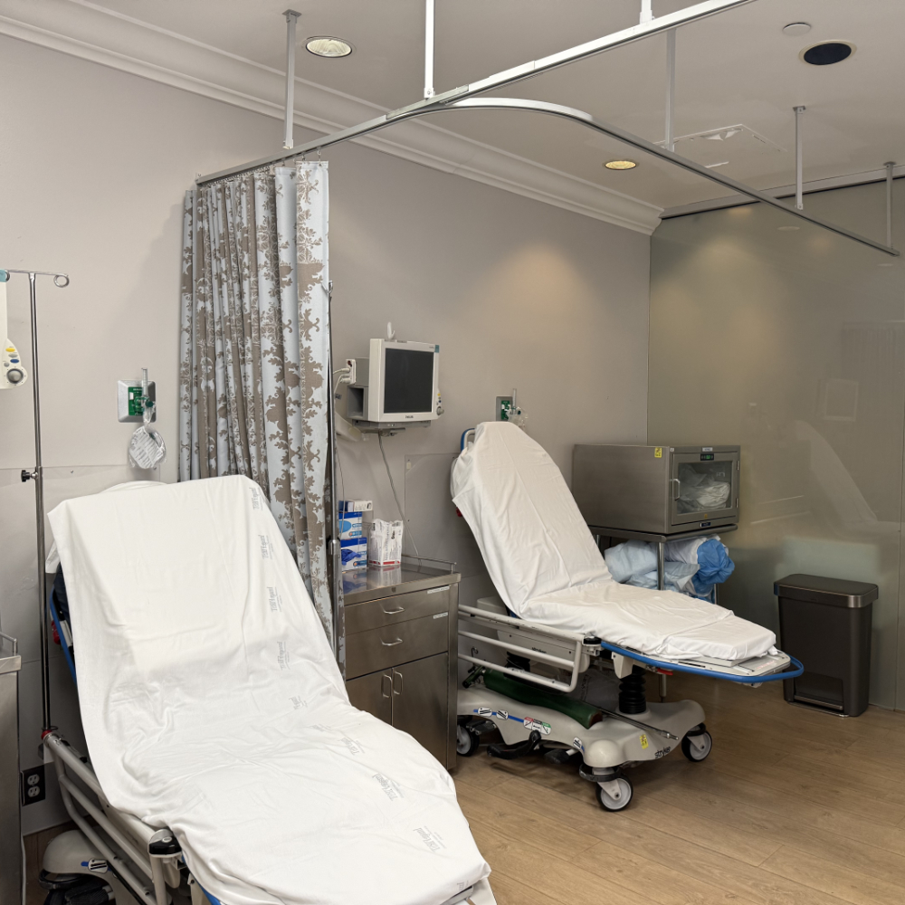 Two empty hospital beds with white sheets in a clean, modern treatment room. Medical equipment, monitors, and a privacy curtain are visible.