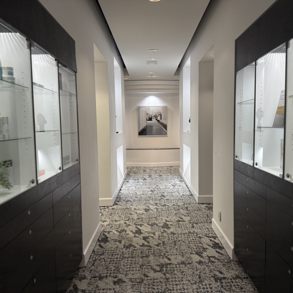 A hallway with glass display cabinets on both sides, patterned carpet, white walls, and a framed photo at the end illuminated by lights.