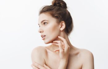 Close up portrait of young female brunette student girl with bun hairstyle and baked shoulders looking aside with calm face expression touching jawline with fingers. Beauty and health